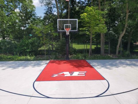 Custom-painted red key area with Appleton East logo on a residential basketball court surrounded by trees